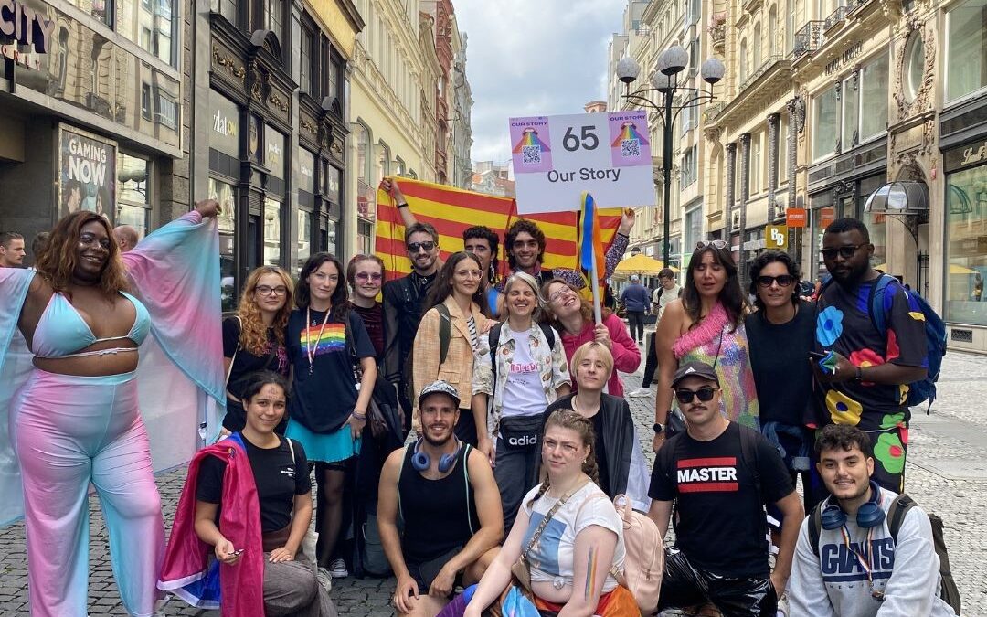 A group of people are standing and smiling while holding the gay and lesbian pride flag. Some are wearing feathers, feather boas, colorful clothing, and are smiling.