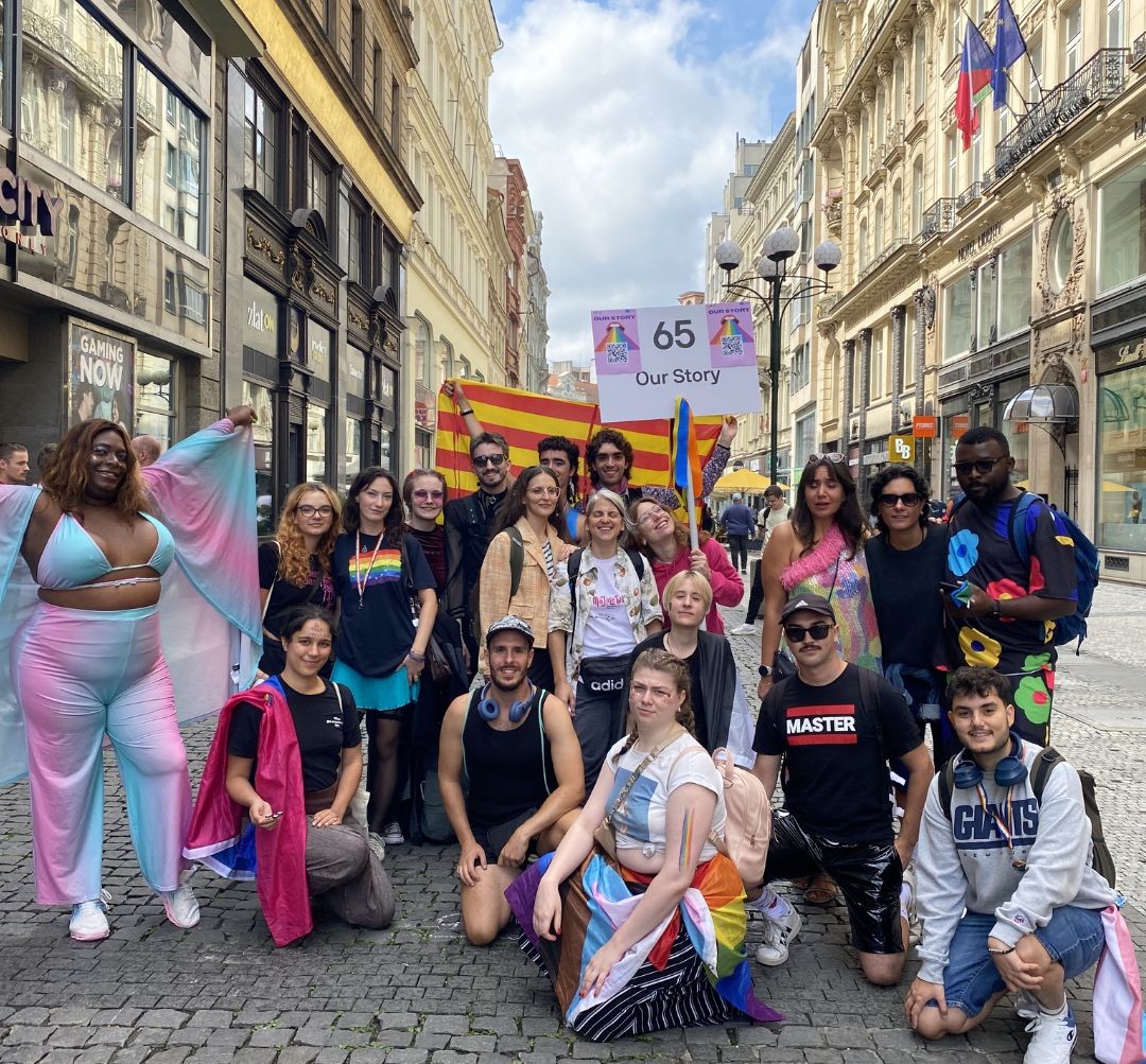 A group of people are standing and smiling while holding the gay and lesbian pride flag. Some are wearing feathers, feather boas, colorful clothing, and are smiling.